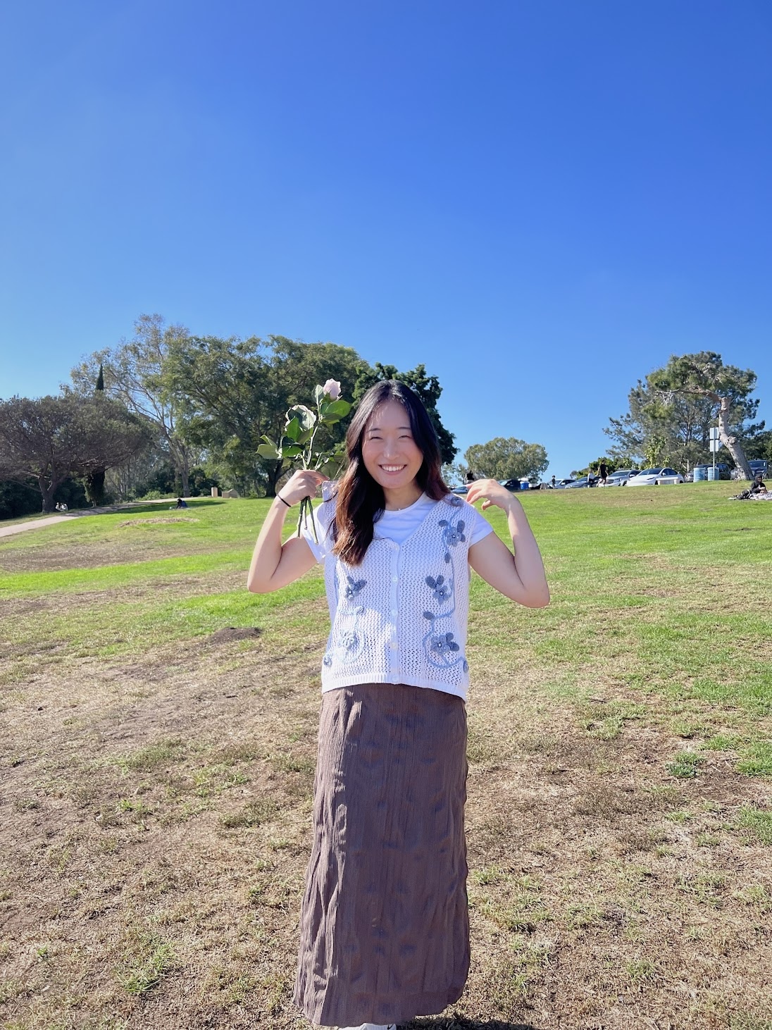 Kate outdoors in a park holding a flower, blue sky behind